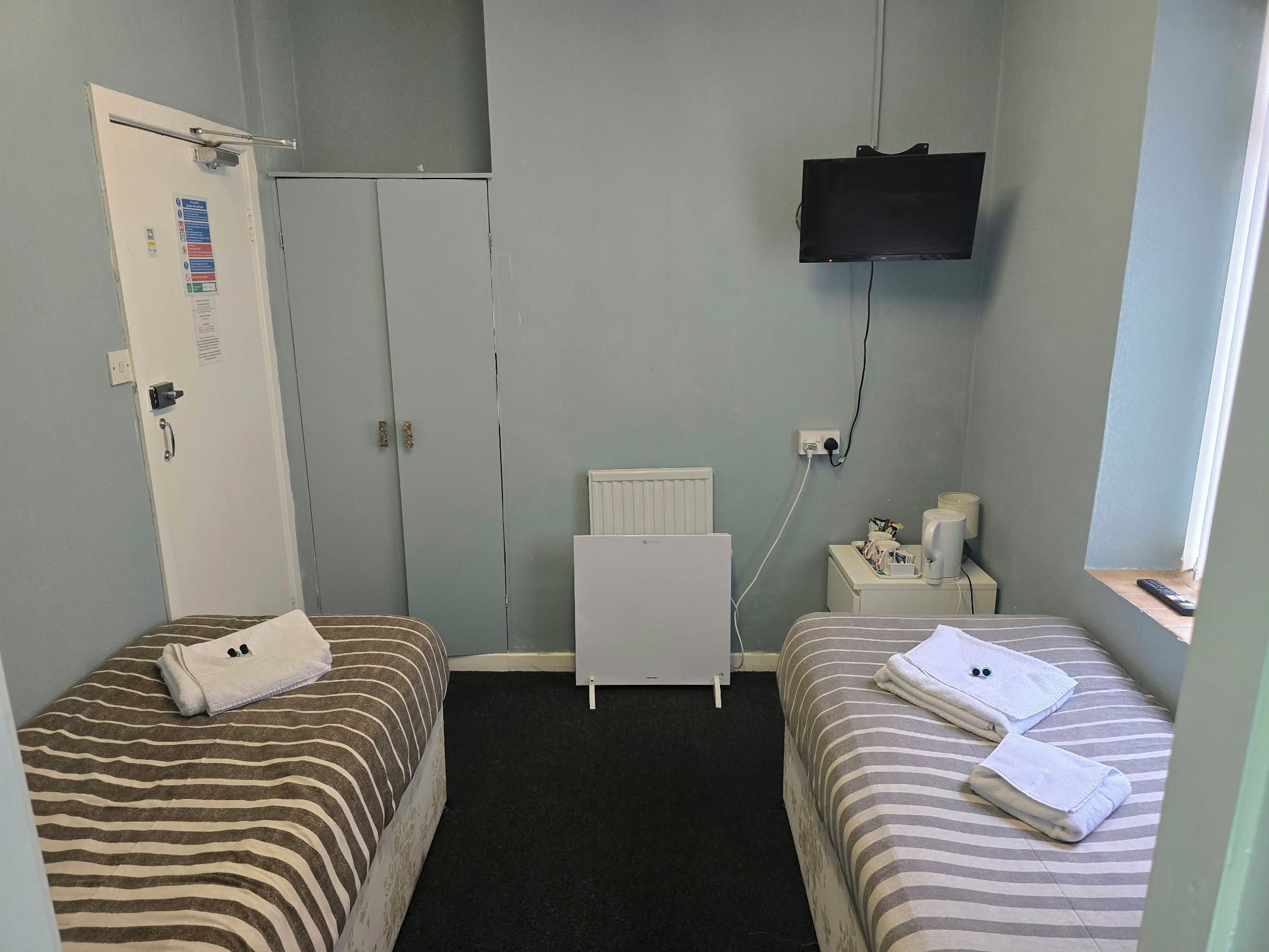 A small hotel room with two beds, a television mounted on the wall, a radiator, and a bedside table with a lamp and a cup.