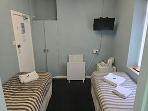 A small hotel room with two beds, a television mounted on the wall, a radiator, and a bedside table with a lamp and a cup. - charnley gold hotel (Blackpool)