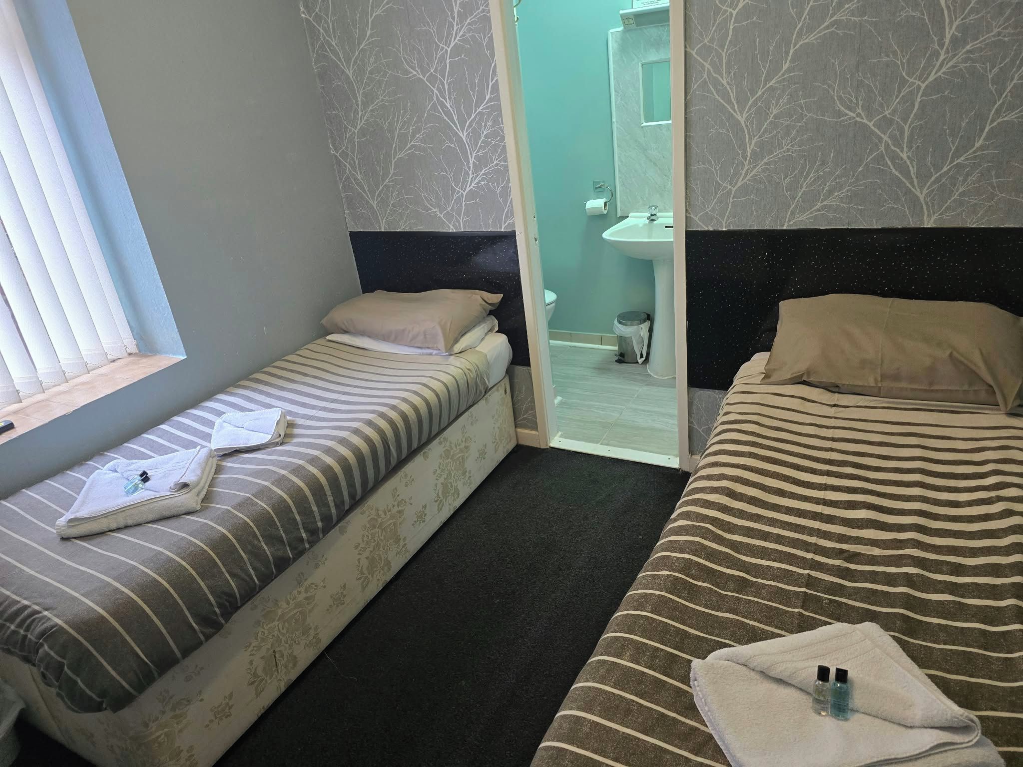 A hotel room with two beds, a window, and a bathroom visible through a glass door.