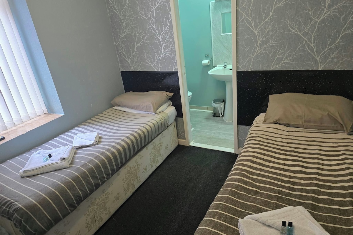 A hotel room with two beds, a window, and a bathroom visible through a glass door.