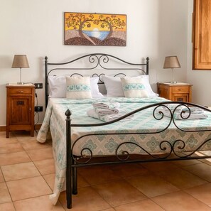 A neatly made bed with a patterned bedspread, two bedside tables with lamps, a wall-mounted painting, and a wooden wardrobe. - Domo Juanna - Cozy Apartment in Laconi (Laconi)
