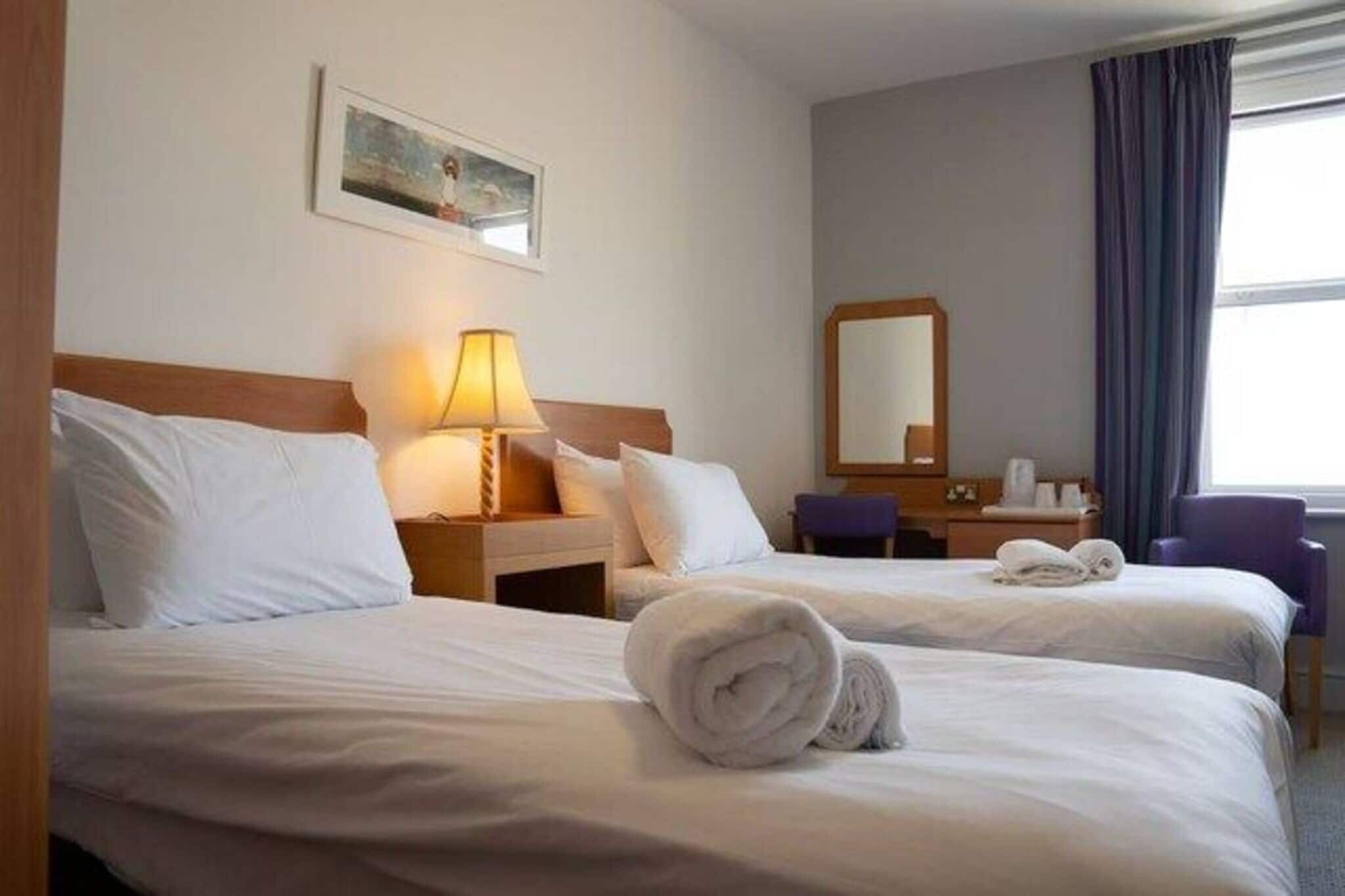 A hotel room with two beds, a desk, a chair, a lamp, a mirror, and a window with curtains.