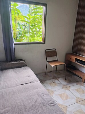 A room with a bed, a chair, a TV stand, and a window with a view of greenery.