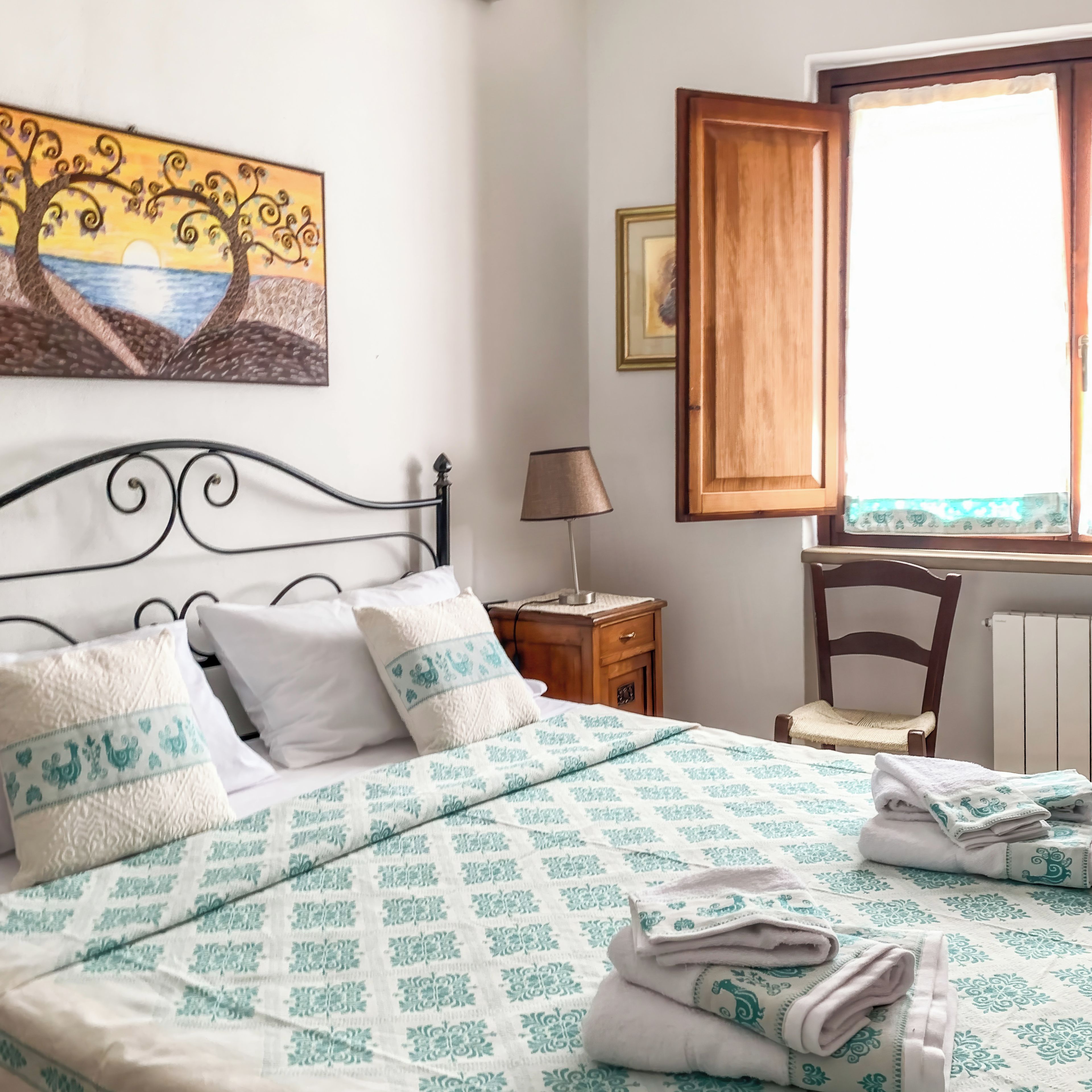 A neatly made bed with a patterned bedspread, pillows, and folded towels. There is a wooden nightstand with a lamp, a chair, and a window with a wooden blind. A painting is visible on the wall.