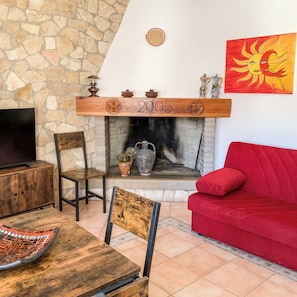 A cozy living room with a stone wall, a fireplace with a year marker, a red sofa, and a television on a wooden cabinet. - Domo Juanna (Laconi)