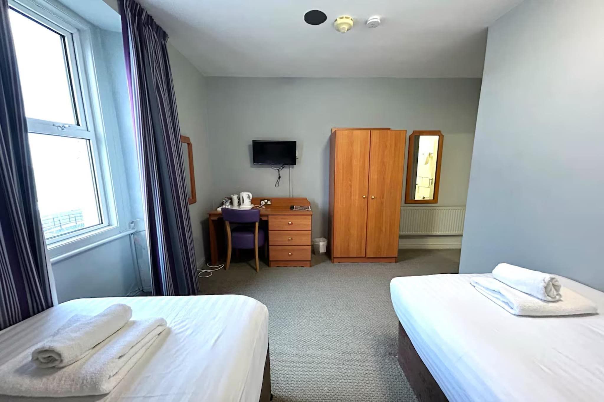 A hotel room with two beds, a desk, a wardrobe, a television, and a window with curtains.