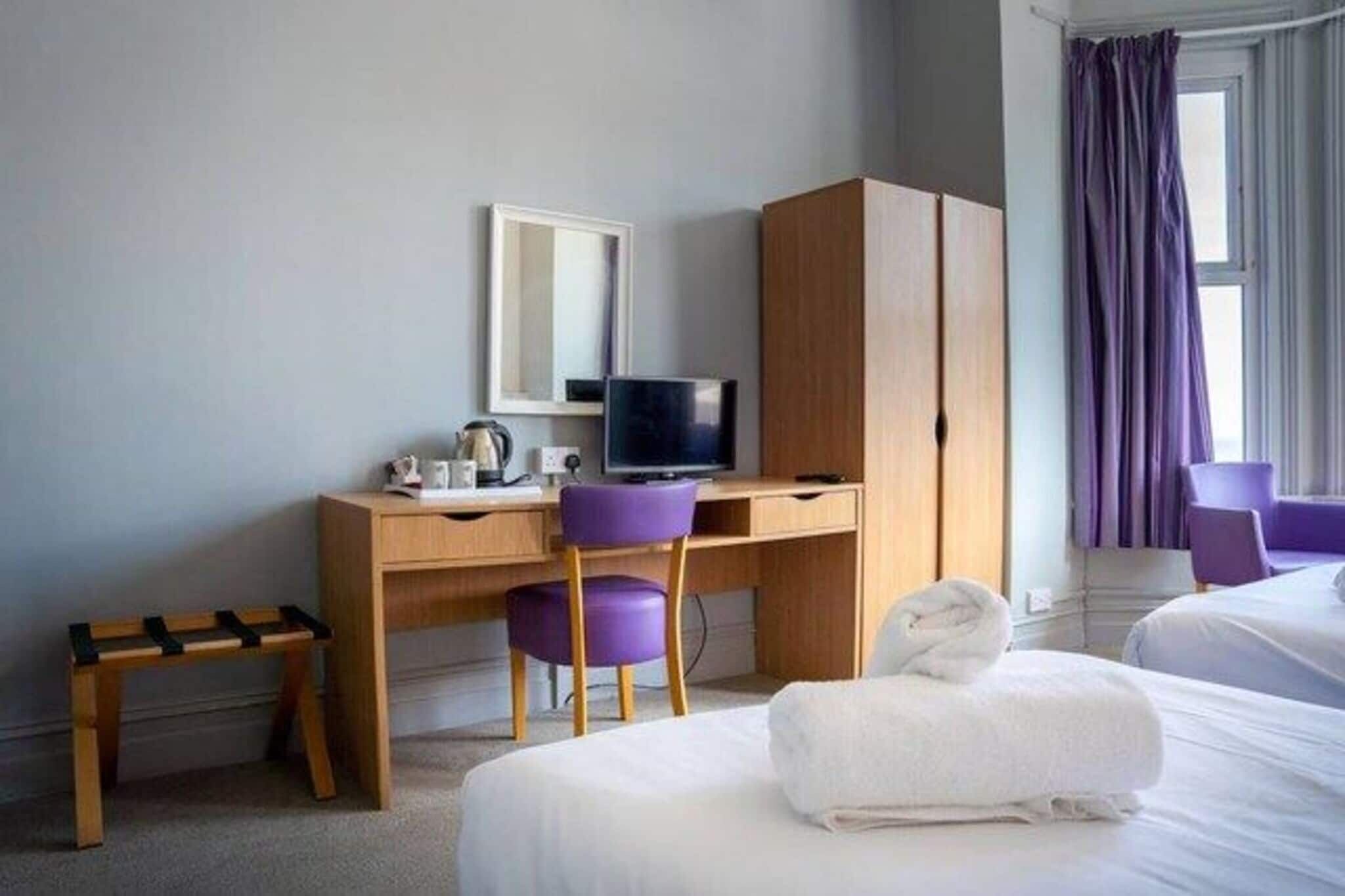 Habitación de hotel con escritorio, silla, televisor, armario, cama y ventana con cortinas.