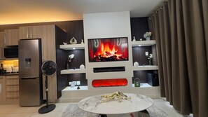 Ein modernes Wohnzimmer mit Flachbildfernseher, integriertem Kamin, Essbereich mit Marmortisch und einer Küche inklusive Kühlschrank.