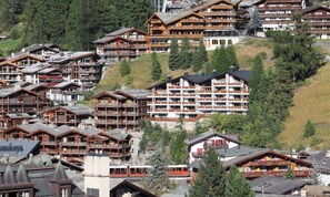A cluster of wooden chalet-style buildings with balconies, set against a backdrop of trees and a hill. - CENTRAL ZERMATT PENTHOUSE WITH SUNNY BALCONY & MATTERHORN VIEWS (Zermatt)