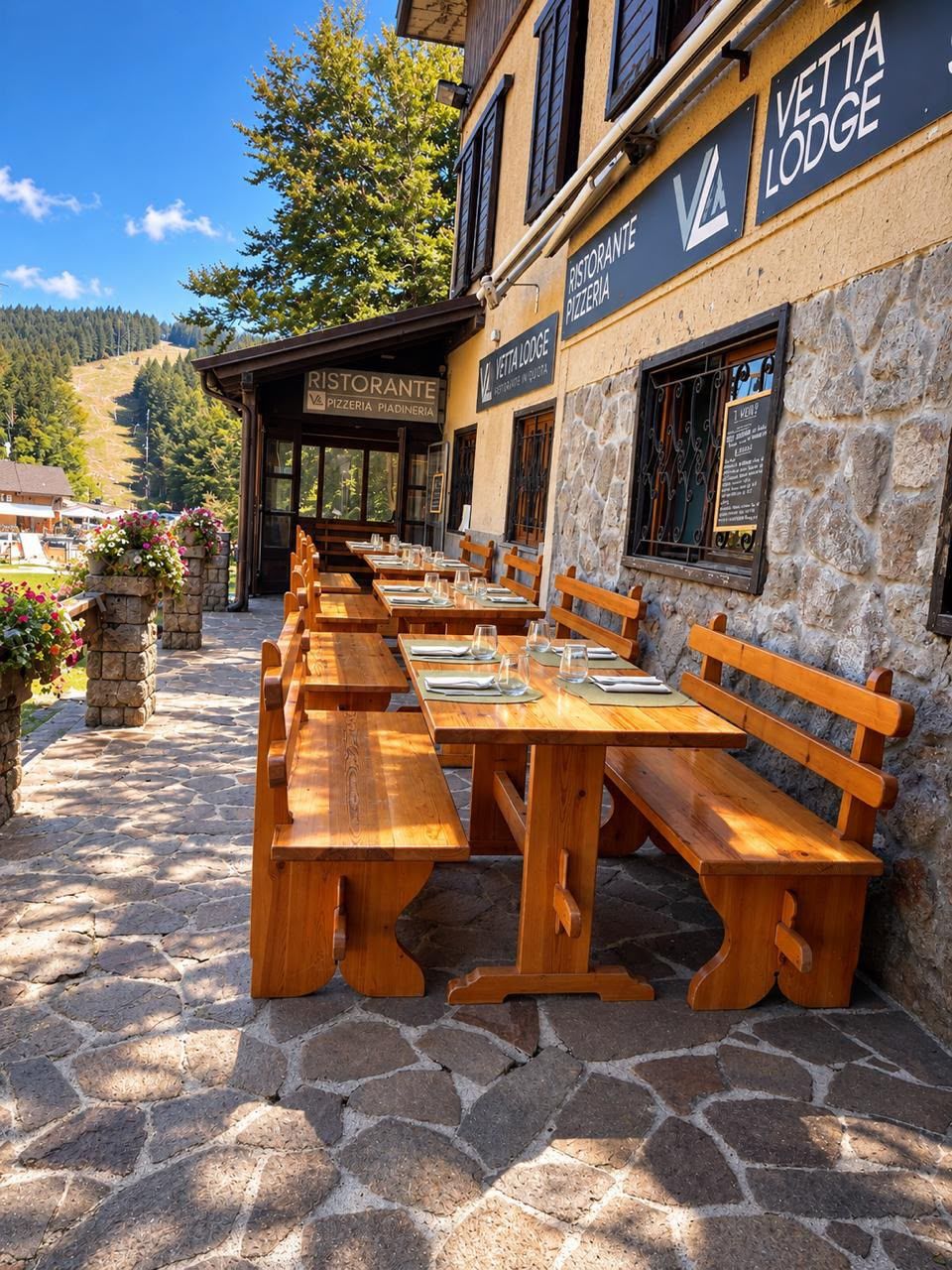 An outdoor seating area with wooden benches and tables, stone flooring, and a building with signage for a restaurant and lodging.