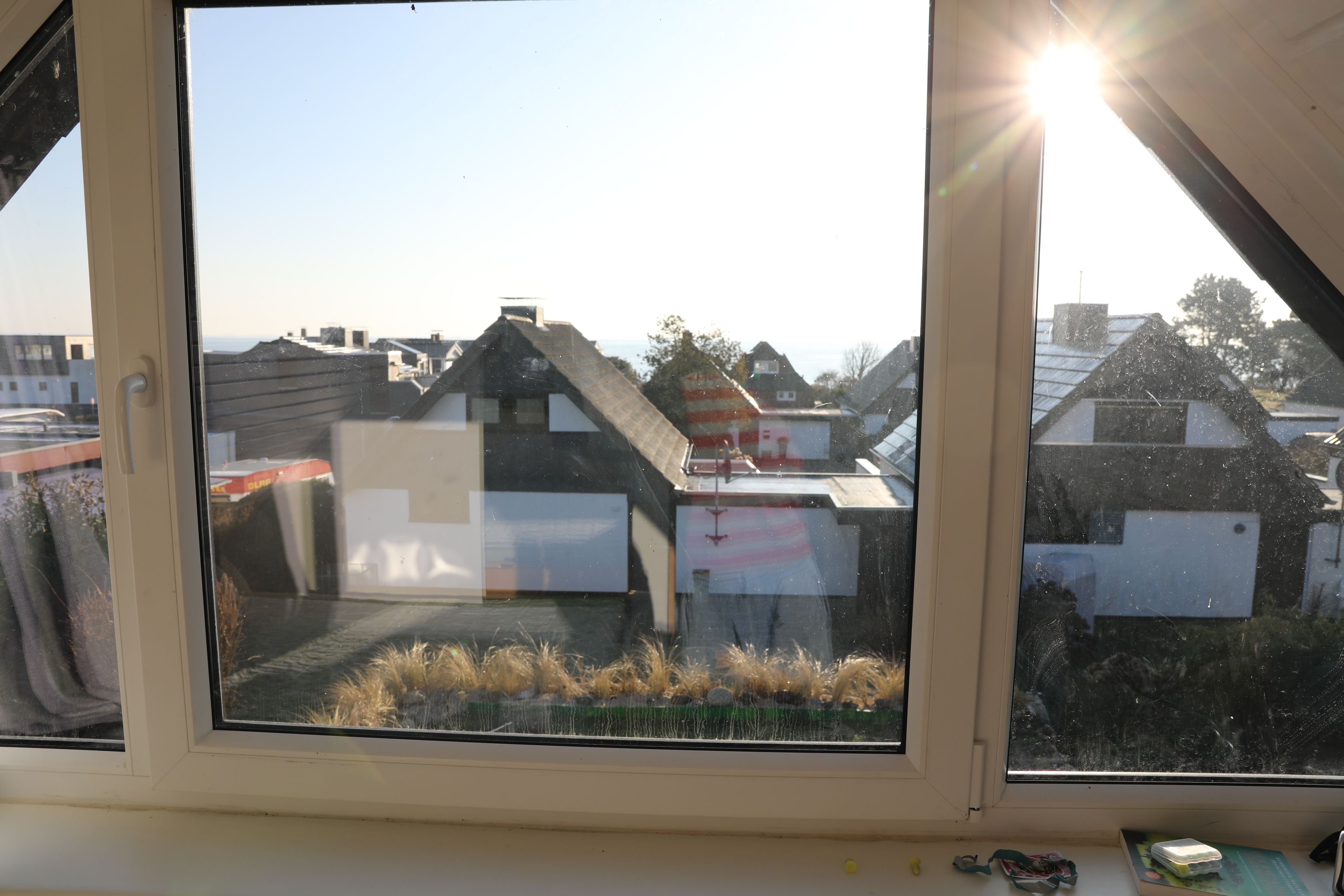 Blick aus einem Fenster bei blauem Himmel, der einen Wohnbereich mit Häusern und ein Gewässer in der Ferne zeigt.