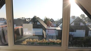 Blick aus einem Fenster bei blauem Himmel, der einen Wohnbereich mit Häusern und ein Gewässer in der Ferne zeigt.