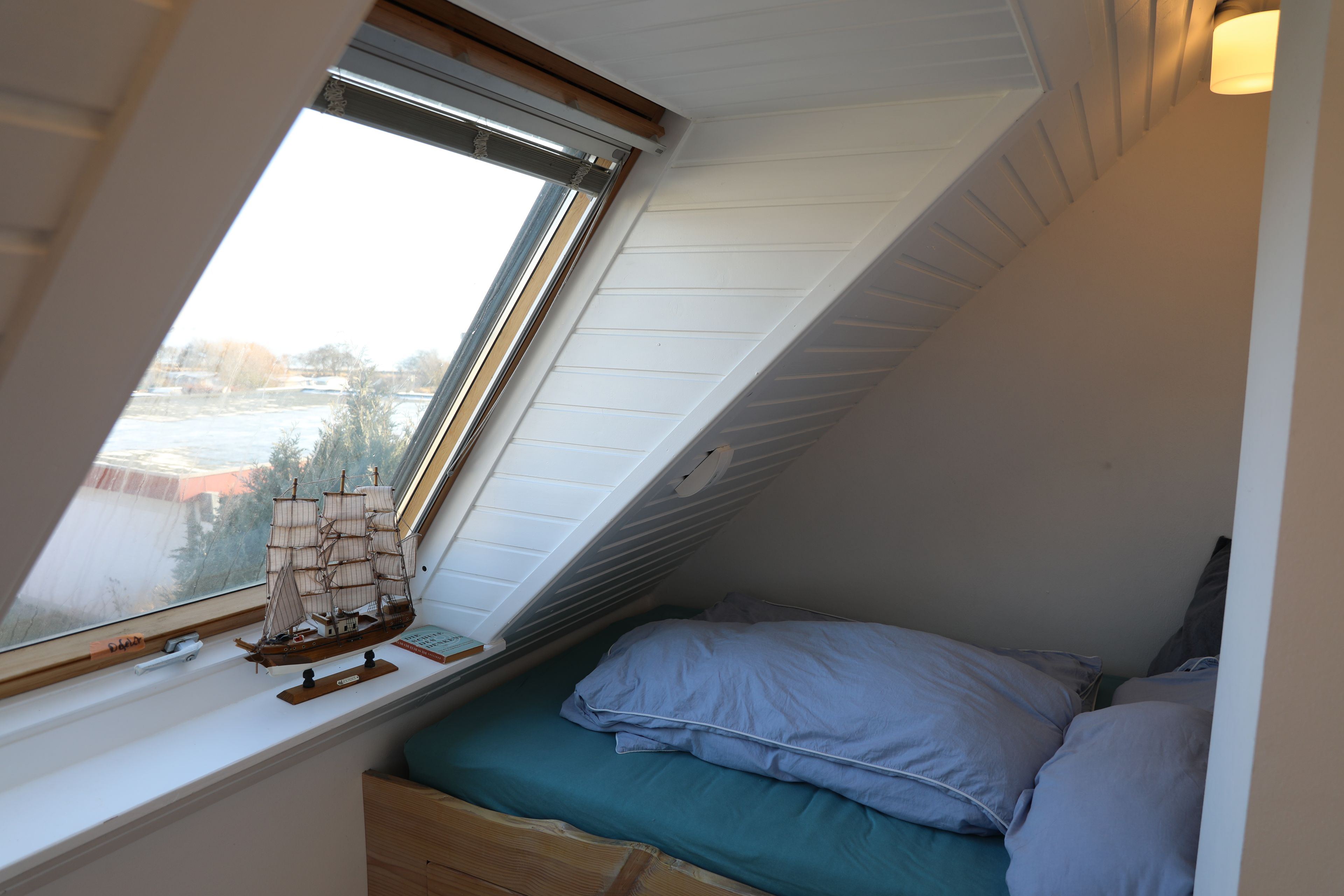 Ein Zimmer mit schrägem Dach, einem Fenster mit Blick auf Bäume und Gebäude, einem Bett mit Kissen und einem Modellschiff auf der Fensterbank.