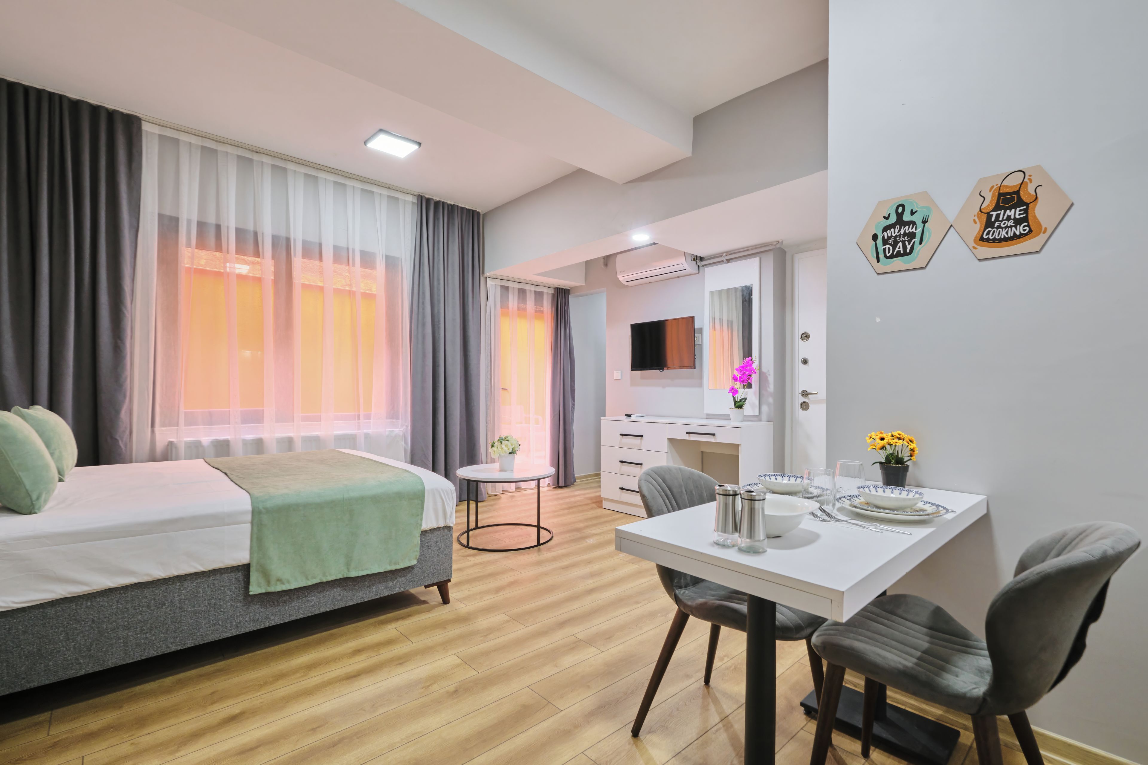 A hotel room with a bed, a dining table with chairs, a small table with a vase, and a mirror.