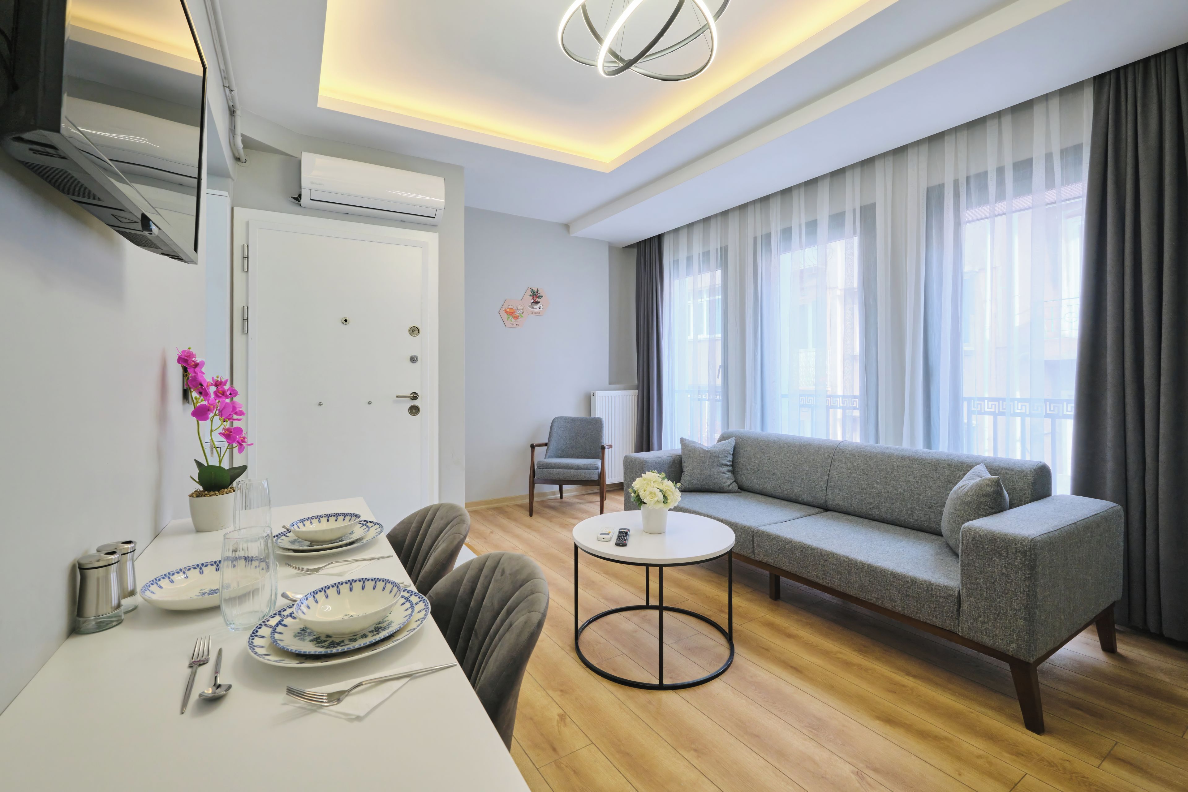 A modern living room with a sofa, a dining table set with plates and cutlery, a small round table with a vase, and a large window with curtains.