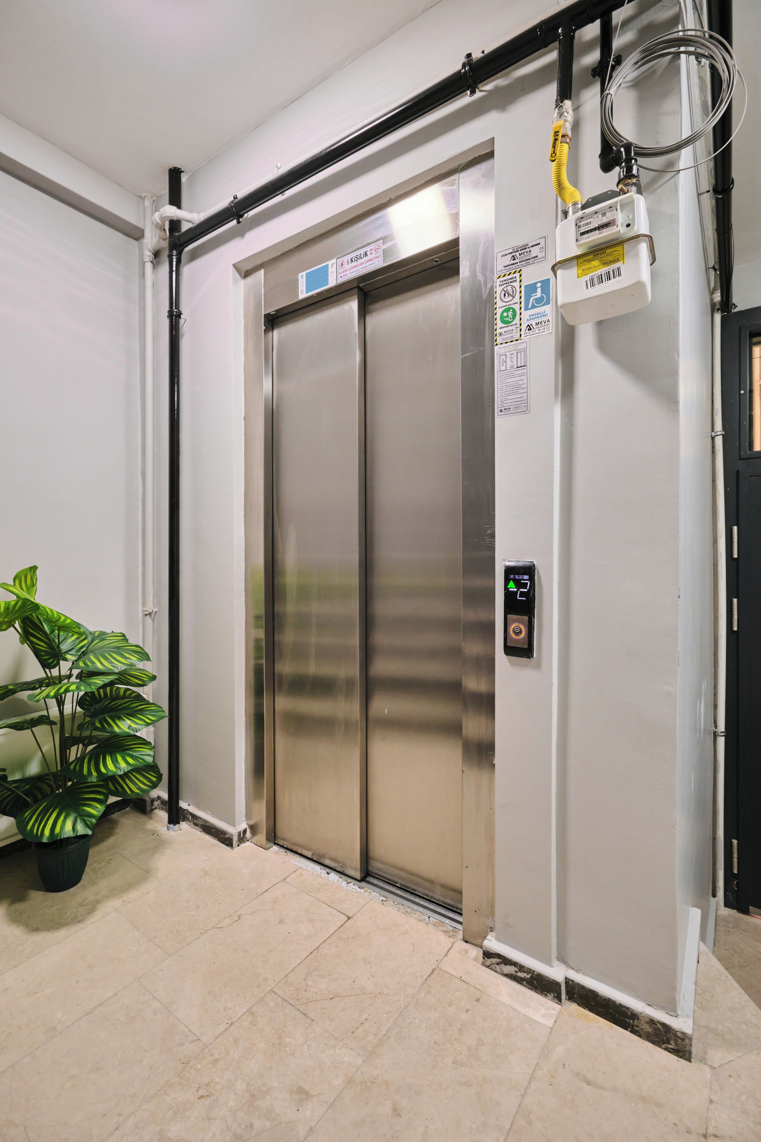 A stationary elevator with a control panel, safety signs, and a potted plant beside it.