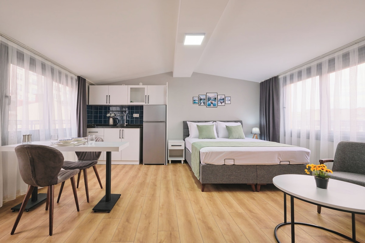 A compact hotel room with a kitchenette, a bed with a green bedspread, a dining table with chairs, and a small round table with a flower vase.