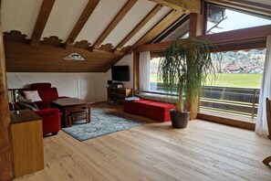 A cozy living room with wooden beams, a red sofa, a coffee table, a TV, and a large window with a view of a village. - Holiday apartment with 2 bedrooms (Bad Hindelang)