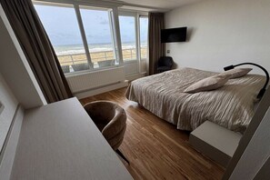 Een hotelkamer met een bed, een bureau, een stoel, een televisie en uitzicht op het strand.