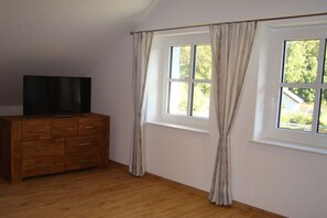Ein Zimmer mit einem Holzkoffer, einem Flachbildfernseher und zwei mit Vorhängen versehenen Fenstern.