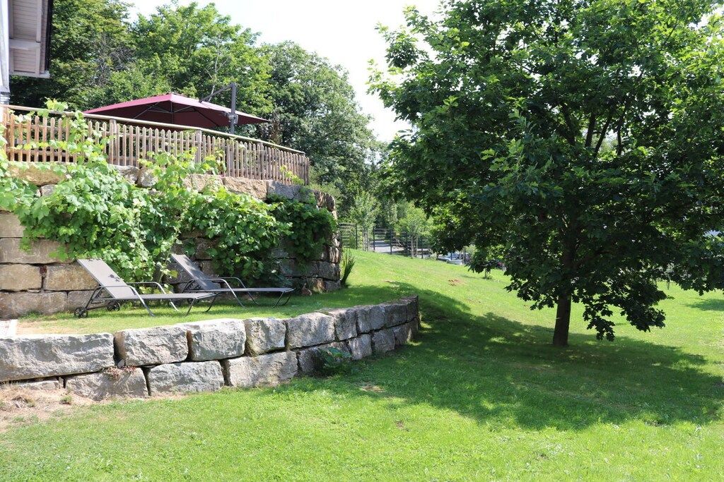 A stone retaining wall, a wooden deck with a red roof, and a large tree in a grassy yard.