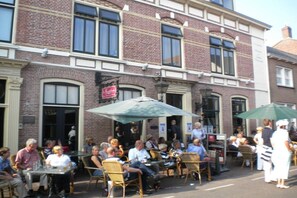 An outdoor cafe with tables and chairs, a brick building with large windows, and a sign above the entrance. - Luxury holiday apartment 2 Zeeland Domburg (Domburg)