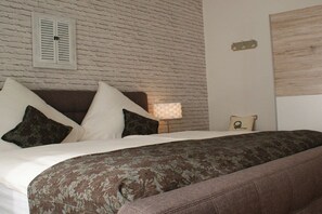 Een slaapkamer met een bakstenen muur, een bed met witte en donkere kussens, een bruine bank en een klein nachtkastje met een lamp.