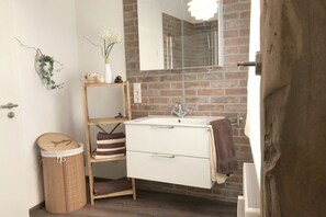 A bathroom with a white sink, a mirror, a wooden shelf with towels, and a bamboo trash bin. - Coach house in the Villa Cues (Benkastel-Kues)