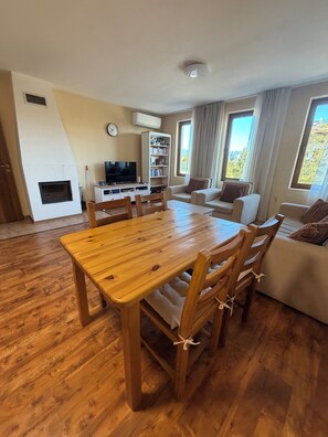 Ampla sala de estar com mesa de jantar de madeira, sofás, televisão e estante de livros.