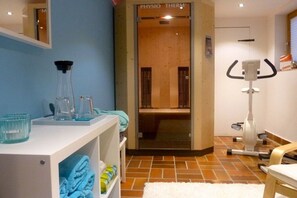 A room with a white shelf holding towels, a glass container, and a water bottle. There's a wooden door labeled 'PHYSIO THERM' leading to a bathroom. A white exercise machine is visible on the right. - Holiday apartment with 0 bedrooms (Reit im Winkl)