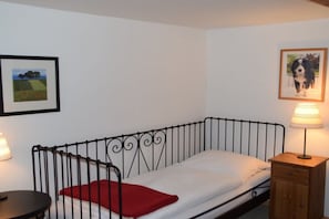 A single bed with a metal frame, a red pillow, a bedside table with a lamp, and two framed pictures on the wall. - Country House Jäger Holiday Apartment No. 2 (Todtnauberg)