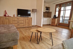 A modern living room with a flat-screen TV, wooden furniture, a small kitchen area, and a balcony with a view. - Holiday apartment with 1 bedroom (Bad Hindelang)