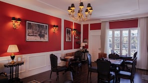 Una habitación con paredes rojas, un candelabro, una mesa de comedor con sillas y un ventanal con cortinas.