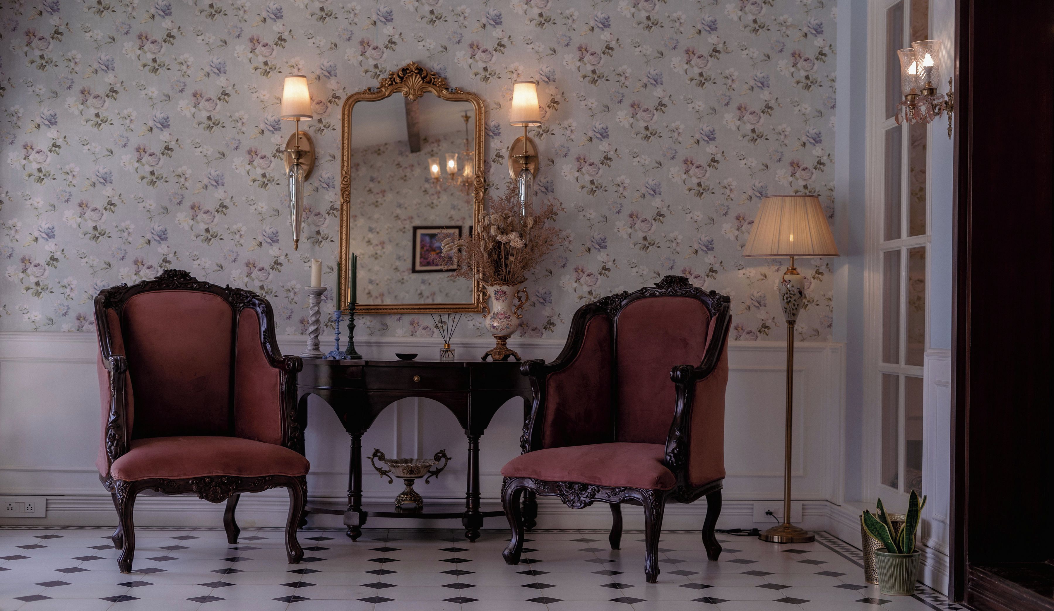 A room with two ornate chairs, a mirror, a table with a vase, and a floor lamp.