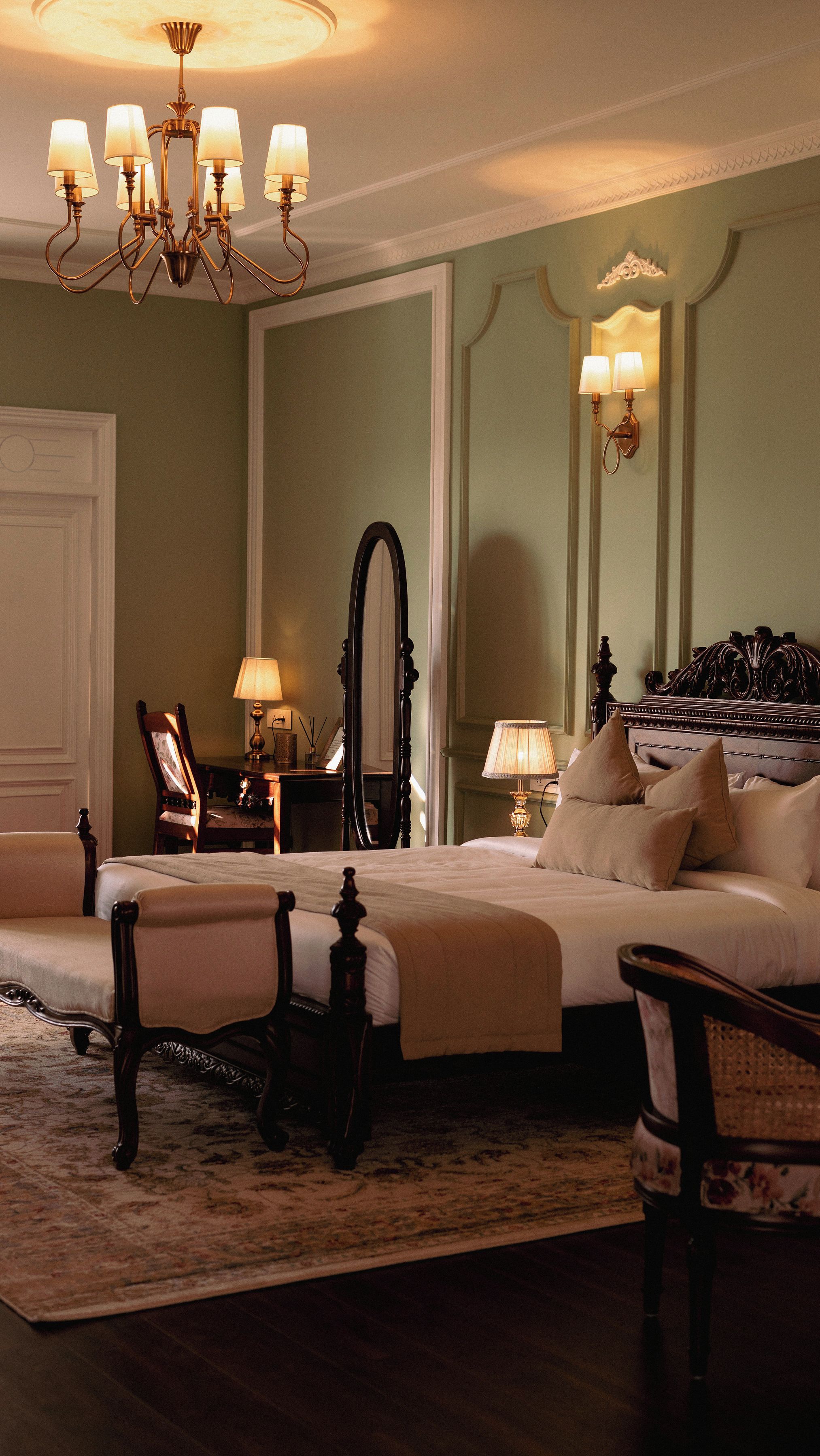 A bedroom with a large bed, a sitting area with a chair, a desk with a lamp, a mirror, and a chandelier.