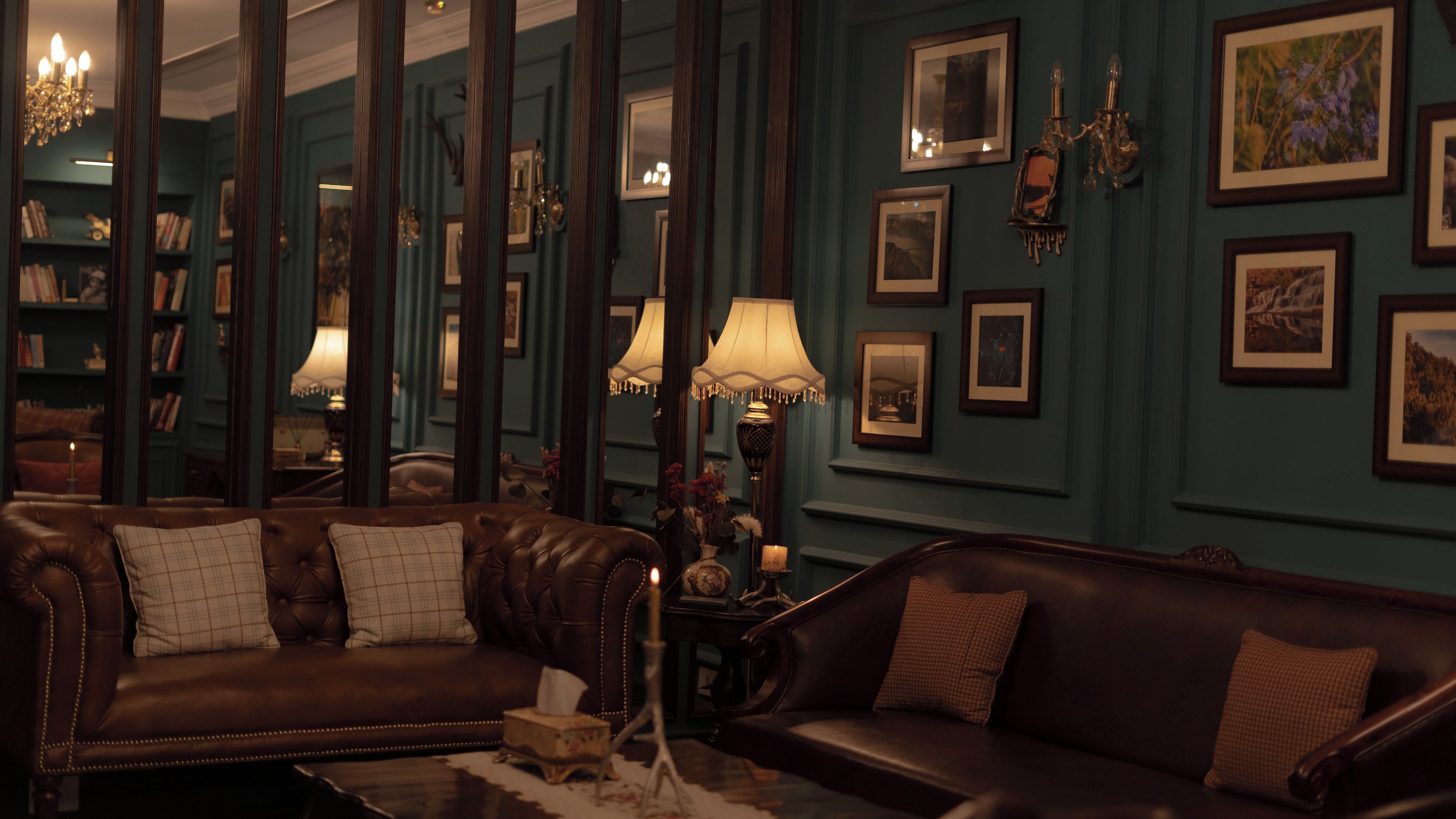 A room with two leather sofas, a chandelier, framed pictures on the wall, and a bookshelf.