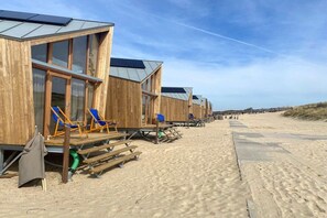 Houten strandhutten met grote ramen en een zandstrand op de achtergrond.