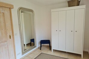 Een kamer met een houten deur, een grote spiegel, een witte kledingkast, een klein bankje en een betegelde vloer.