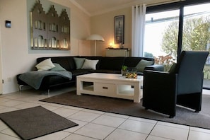A living room with a black sofa, a white coffee table, and a black armchair. There is a radiator, a lamp, and a window with a view of trees. - Bungalow in Lemmer with swimming pool (Lemmer)
