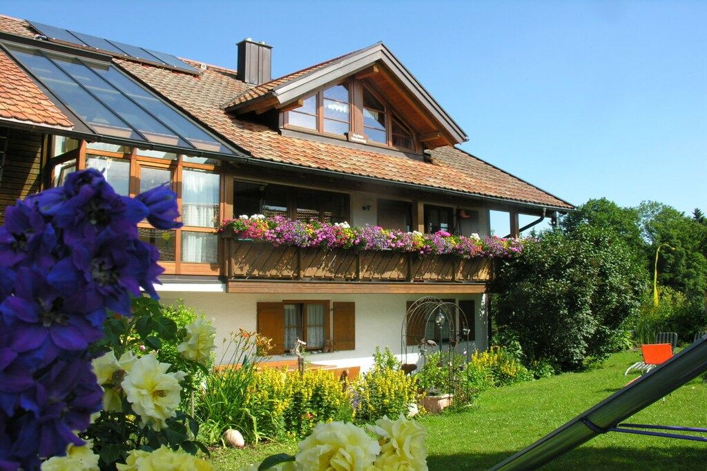 A two-story house with a sloped roof, large windows, and a balcony with flowers.
