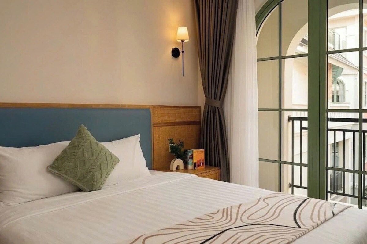Una habitación de hotel con cama, mesita de noche, lámpara fijada a la pared y vistas por un ventanal grande.