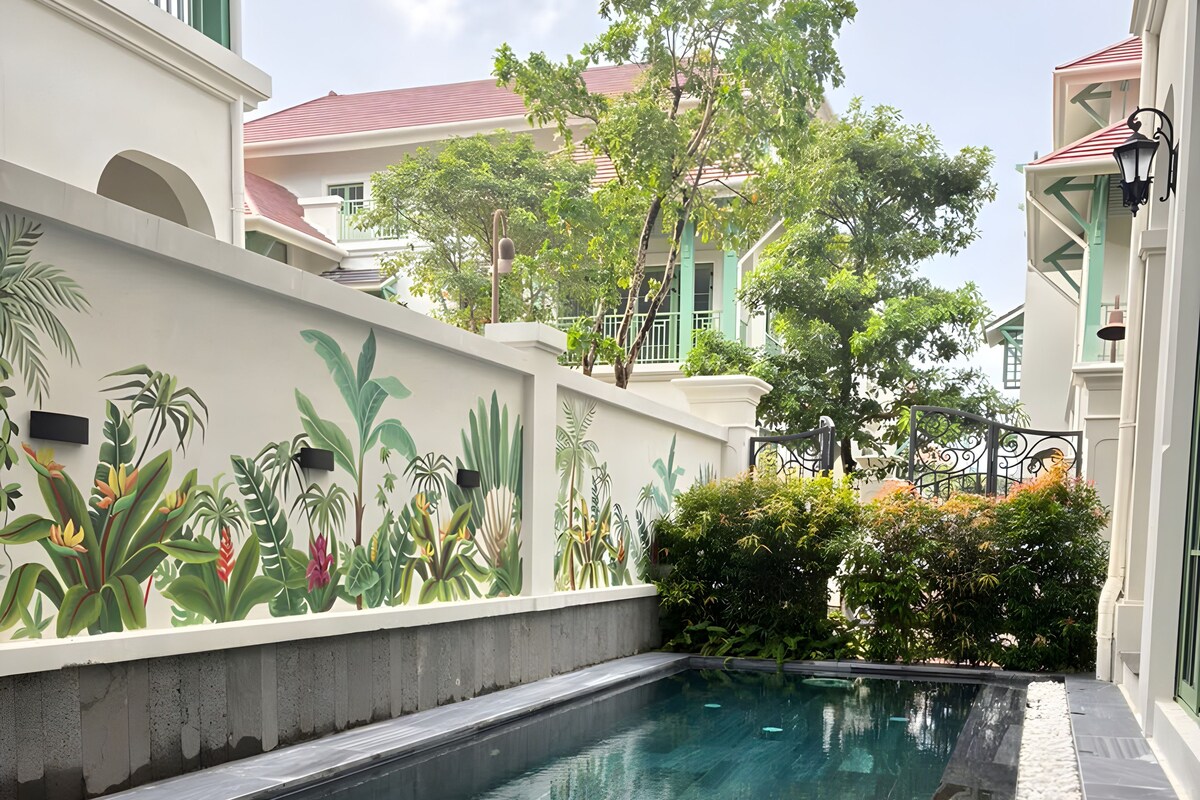 Una piscina con superficie de agua cristalina, rodeada por un muro bajo con un mural de plantas tropicales, y edificios colindantes con balcones.