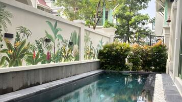 Una piscina con superficie de agua cristalina, rodeada por un muro bajo con un mural de plantas tropicales, y edificios colindantes con balcones.
