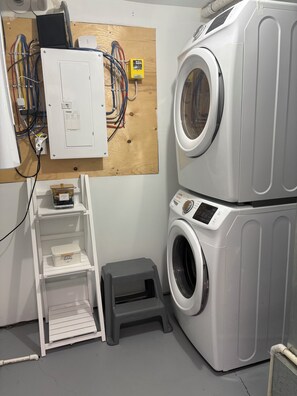 Ein Waschraum mit nebeneinander gestapelten Waschmaschine und Trockner, einer Holztruhe mit elektrischer Verkabelung und einem kleinen Hocker.