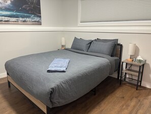 A bedroom with a bed, pillows, a nightstand, a lamp, and a wall-mounted window. - Cozy 2-bedroom, 1-bath Lower Duplex on North Side - Oasis Corporate Rentals (Grande Prairie)