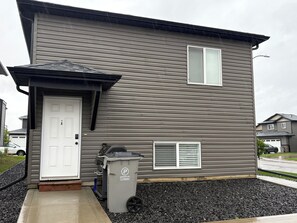 A two-story house with a grey siding exterior, a white front door, a small attached garage, and a trash bin with a recycling symbol. - Cozy 2-bedroom, 1-bath Lower Duplex on North Side - Oasis Corporate Rentals (Grande Prairie)