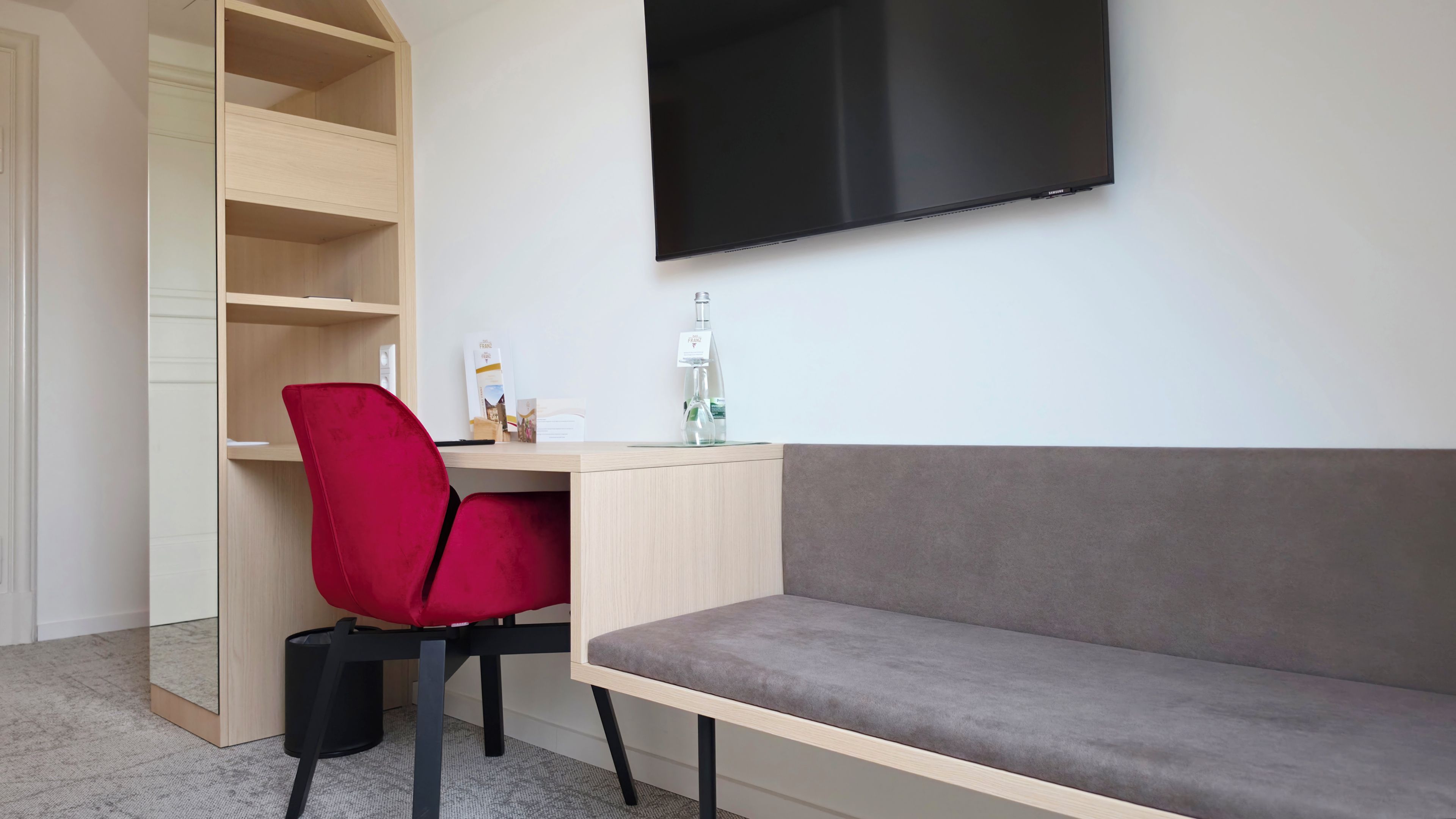 Ein modernes Zimmer mit Schreibtisch, einem roten Sessel, einem grauen Sofa und einem flachbildfernseher an der Wand.