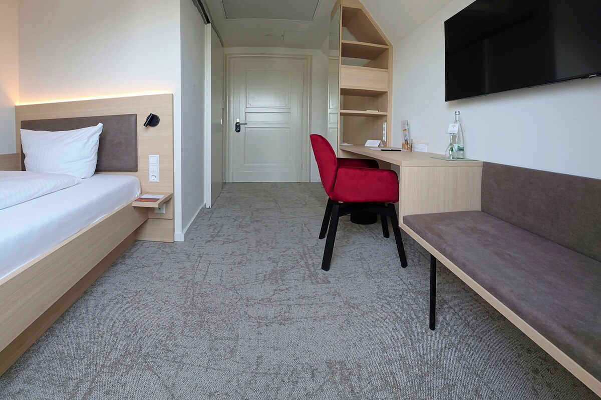 Una camera d'albergo con un letto, una scrivania, una poltrona rossa, un divano e una TV a schermo piatto montata a parete.