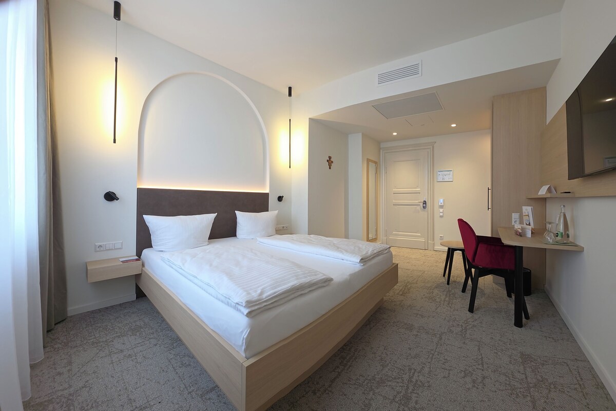 Una camera d'albergo con un letto, comodini, una scrivania con una sedia, una televisione e una porta che dà su un'altra stanza.
