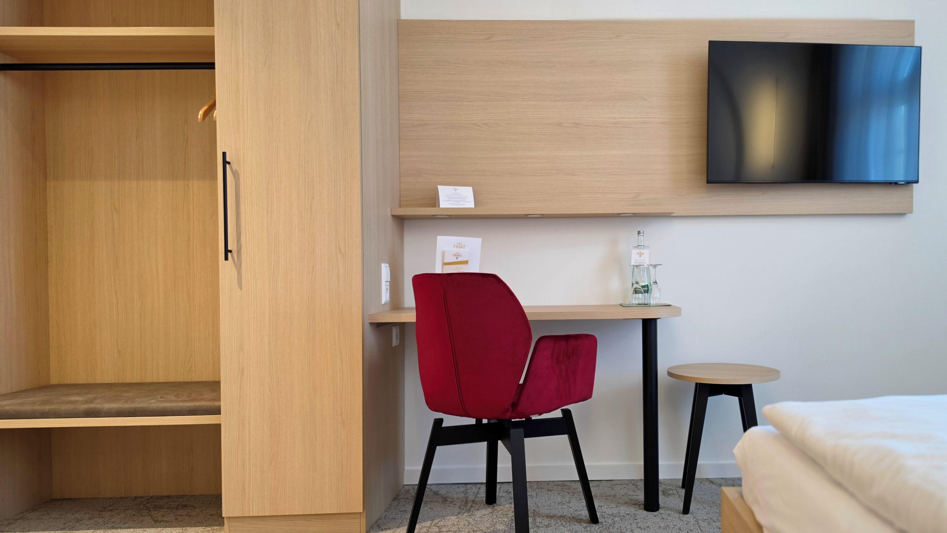 A hotel room with a wooden wardrobe, a red chair, a small desk with a bottle and glasses, a television, and a bed.
